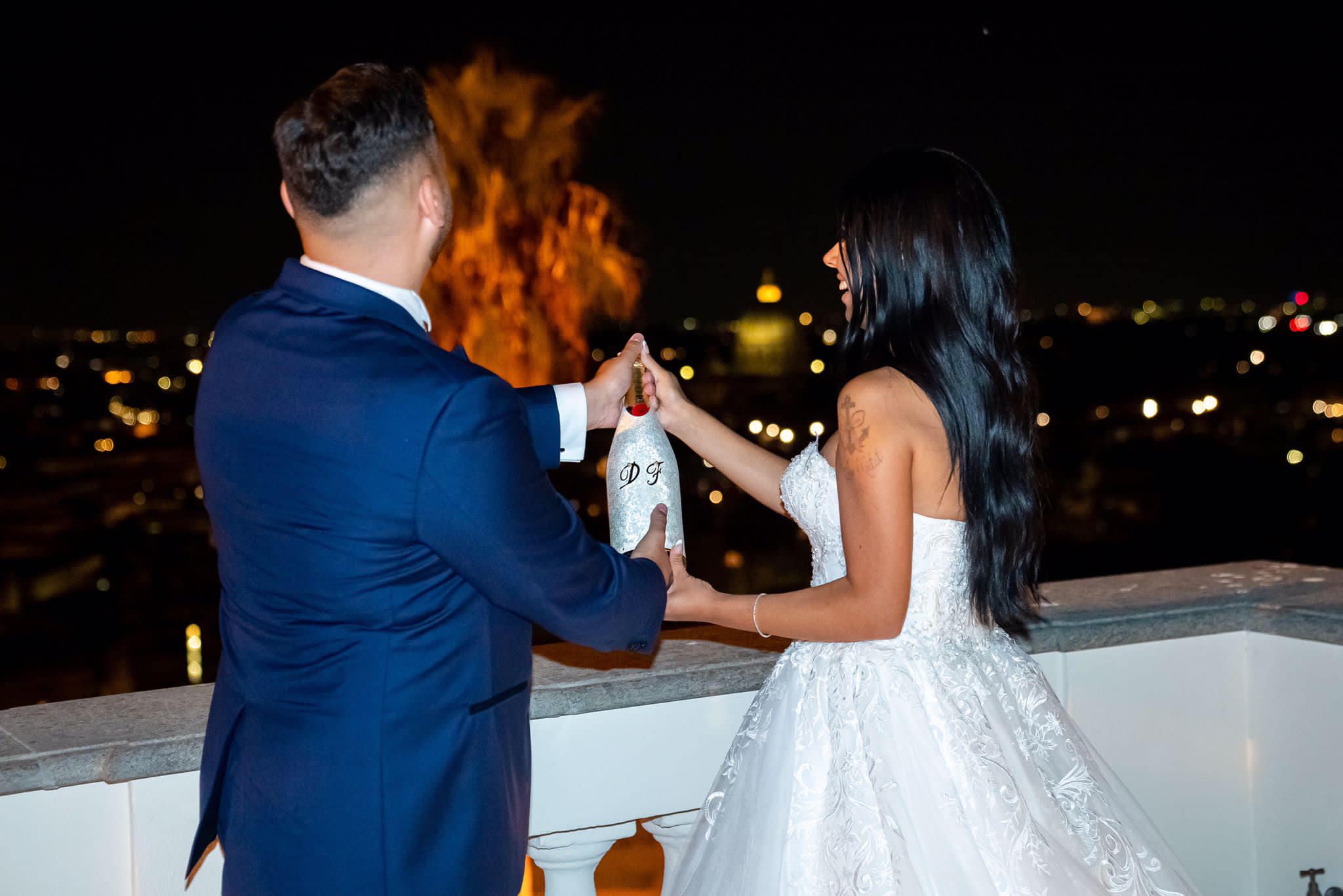 Bride and groom sharing a toast moment, holding a personalized champagne bottle with their initials, against a romantic night skyline.