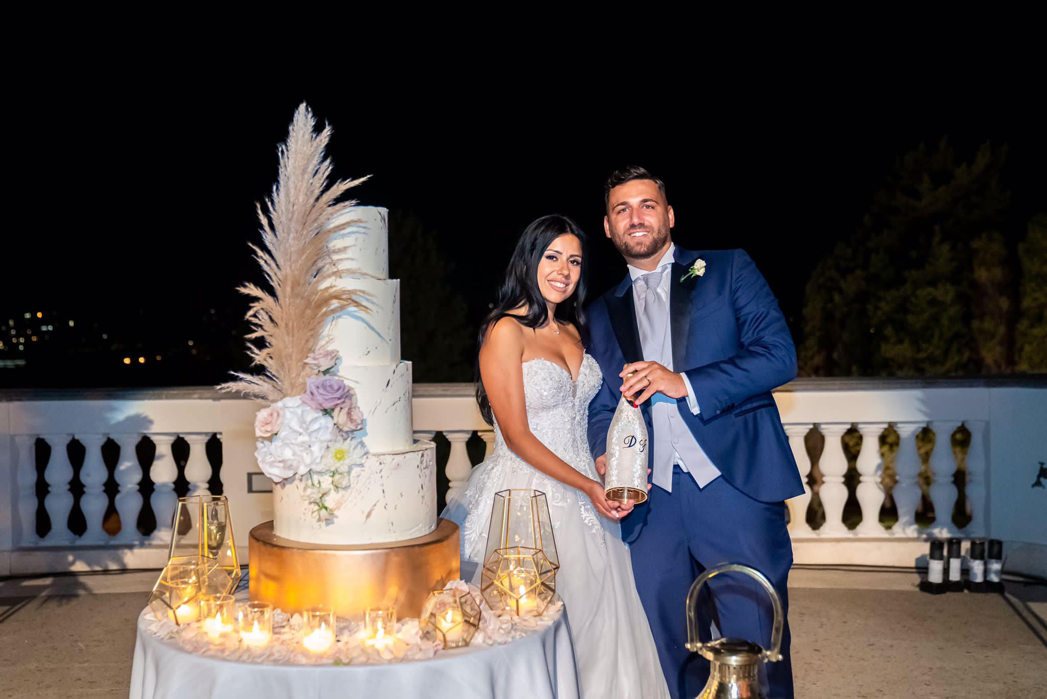 Bride and groom posing by their wedding cake, holding a champagne bottle personalized with their initials and adorned with a silver textured cover.