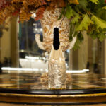 The champagne bottle is dressed with the 'Ariosto' cover, a silver creation inspired by the epic tales of Ludovico Ariosto. The cover features a finely sculpted battle scene between a knight and a dragon, evoking the imagery of Orlando Furioso. Positioned on a richly decorated inlaid table, with a grand vase and autumnal foliage in the background, the setting enhances the sense of art, history and luxury.