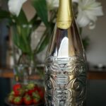 A champagne bottle adorned with a silver cover featuring an elaborate tribal design. The handcrafted cover, made from precious metal, wraps around the lower part of the bottle, transforming it into a decorative art piece. In the background, fresh strawberries and white lilies complete the refined and luxurious atmosphere