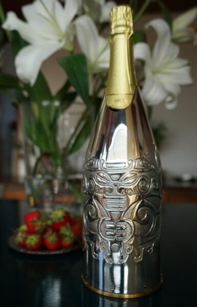 A champagne bottle adorned with a silver cover featuring an elaborate tribal design. The handcrafted cover, made from precious metal, wraps around the lower part of the bottle, transforming it into a decorative art piece. In the background, fresh strawberries and white lilies complete the refined and luxurious atmosphere