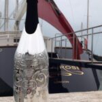 A silver-engraved champagne bottle cover with an intricate tribal design stands on a stone surface in front of a luxury yacht, with a red maritime flag waving in the background under an overcast sky.