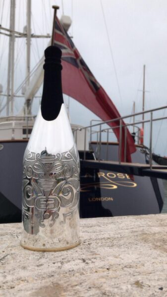 A silver-engraved champagne bottle cover with an intricate tribal design stands on a stone surface in front of a luxury yacht, with a red maritime flag waving in the background under an overcast sky.