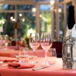 The champagne bottle features the 'Flower Chain' cover, a handcrafted silver design that frames the central part of the bottle with an intricate garland of flowers, leaves, and ribbons. The bottle is placed on an elegant table set for a refined dinner, with pink tablecloths, neatly folded napkins, and crystal glasses, immersed in the soft glow of evening lights.