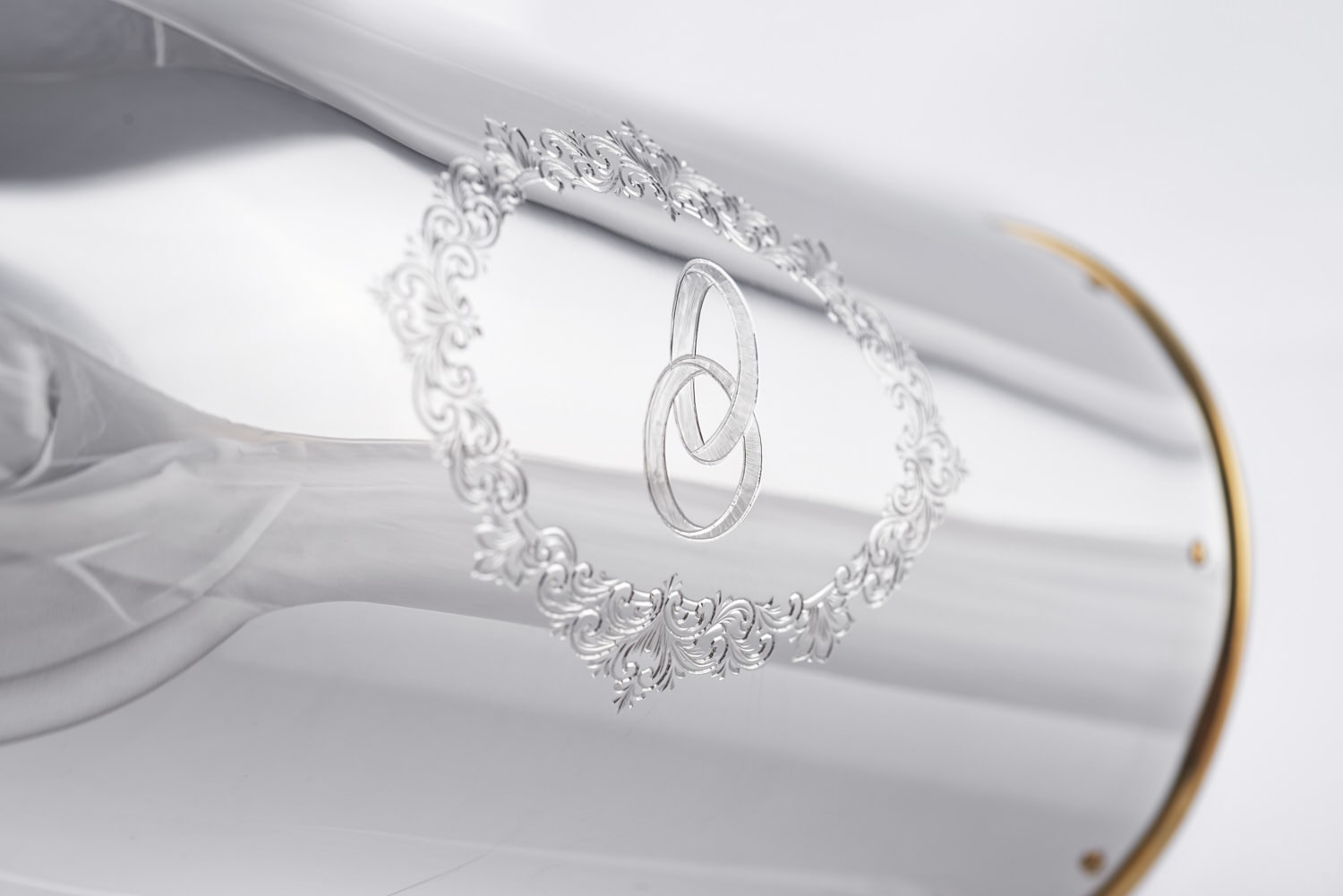 Close-up of the 'Wedding Rings' champagne bottle cover, featuring two interlocking rings engraved within an ornate floral frame on polished silver.