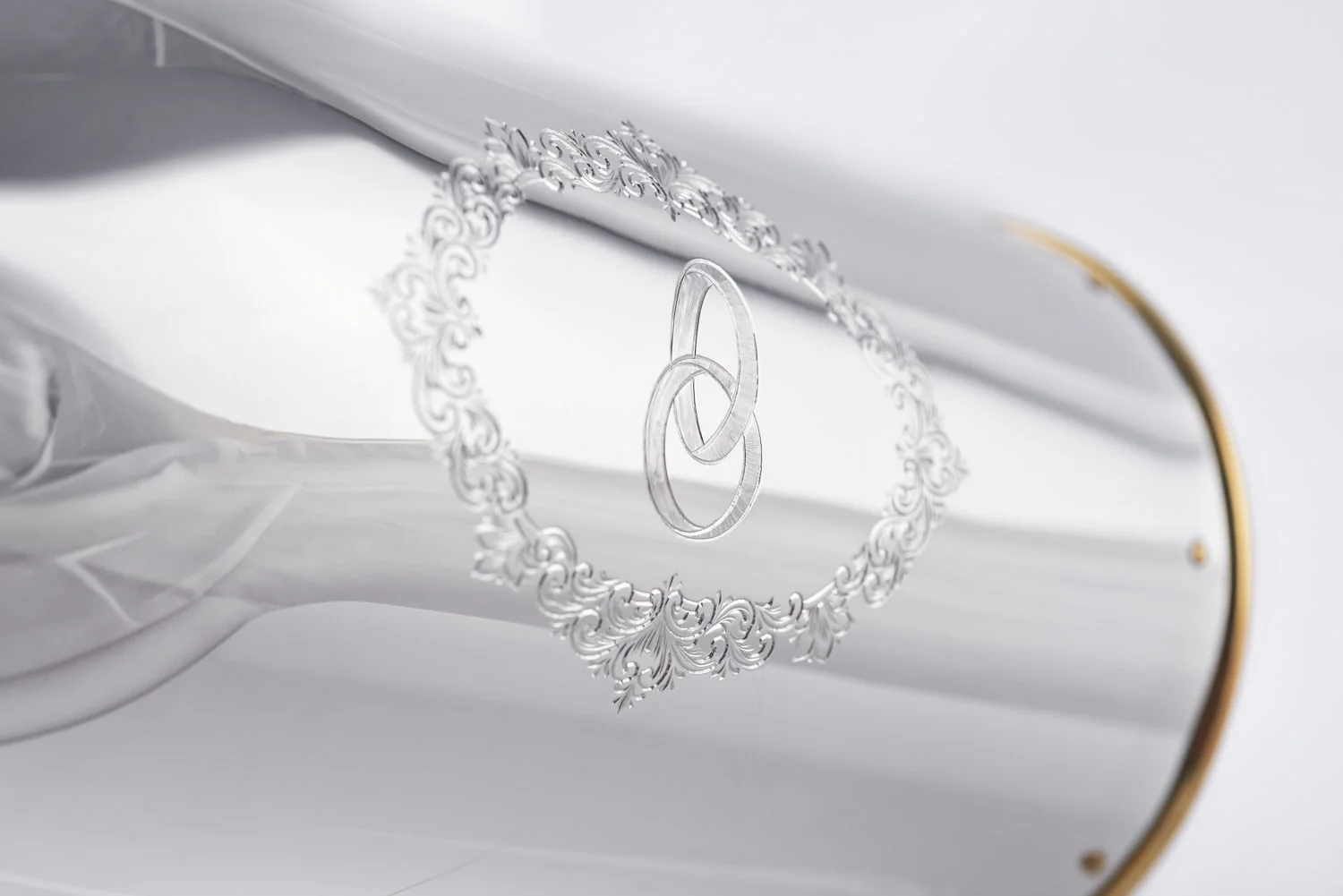 Close-up of the 'Wedding Rings' champagne bottle cover, featuring two interlocking rings engraved within an ornate floral frame on polished silver.